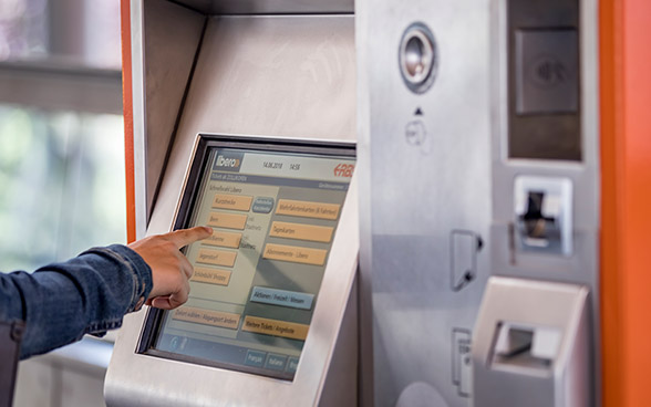 A ticket vending machine with touchscreen, showing a hand in the foreground.